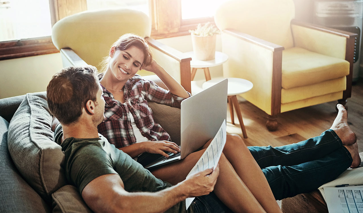 http://Couple%20sits%20in%20their%20living%20room,%20smiling%20at%20one%20antoher%20while%20holding%20financial%20documents